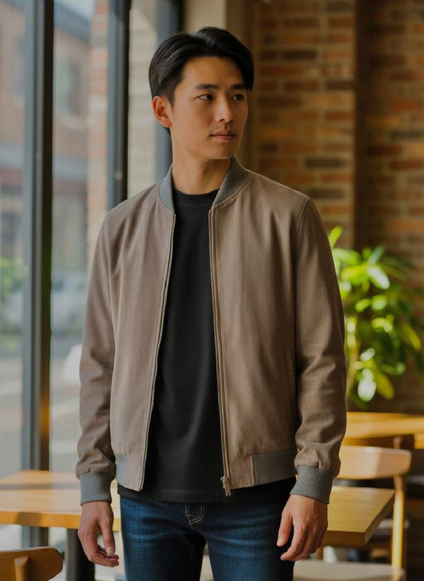 Man wearing stylish beige bomber jacket with gray trim, paired with black tee and jeans in modern cafe setting